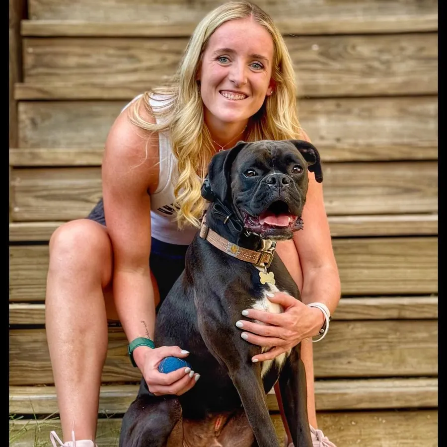 An owner kneeling with her black dog against a wooden fence